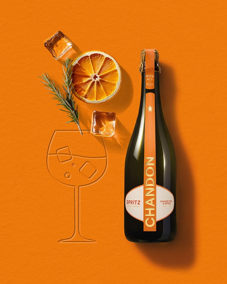 Chandon Spritz bottle, dried orange slice, rosemary sprig, and ice cubes arranged on an orange background next to an embossed outline of a wine glass
