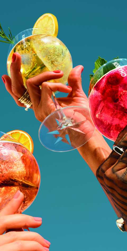 Close-up of three hands holding Chandon Spritz Orange, Lemon and Berries cocktail glasses, garnished with citrus and rosemary, against a vibrant blue sky